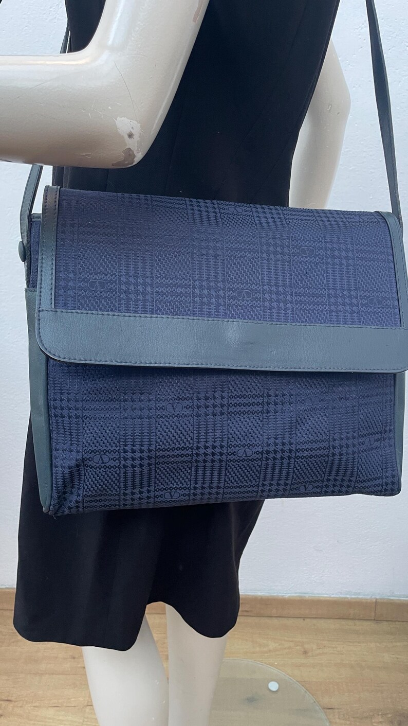May include: A navy blue shoulder bag with a flap closure. The bag features a plaid pattern and a gray leather trim. The strap is also gray. The bag is displayed on a mannequin wearing a black dress.