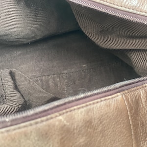 May include: Open view of a brown leather handbag interior. The inside is lined with a brown fabric with a striped pattern. The zipper is visible along the top edge of the bag.