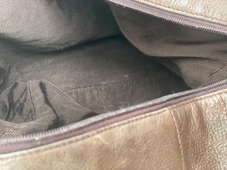 May include: Open view of a brown leather handbag interior. The inside is lined with a brown fabric with a striped pattern. The zipper is visible along the top edge of the bag.