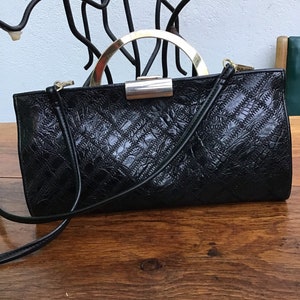 Op de afbeelding: Zwarte rechthoekige handtas met een textuurpatroon en een goudkleurige handgreep. De tas heeft een lange, dunne riem en een zilverkleurige sluiting. De tas staat op een houten oppervlak.