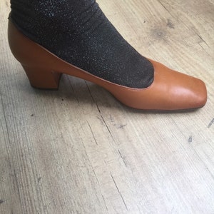 May include: A brown leather shoe with a square toe and a small heel. The shoe is paired with a black, sparkly sock. The shoe is on a light brown wooden floor.