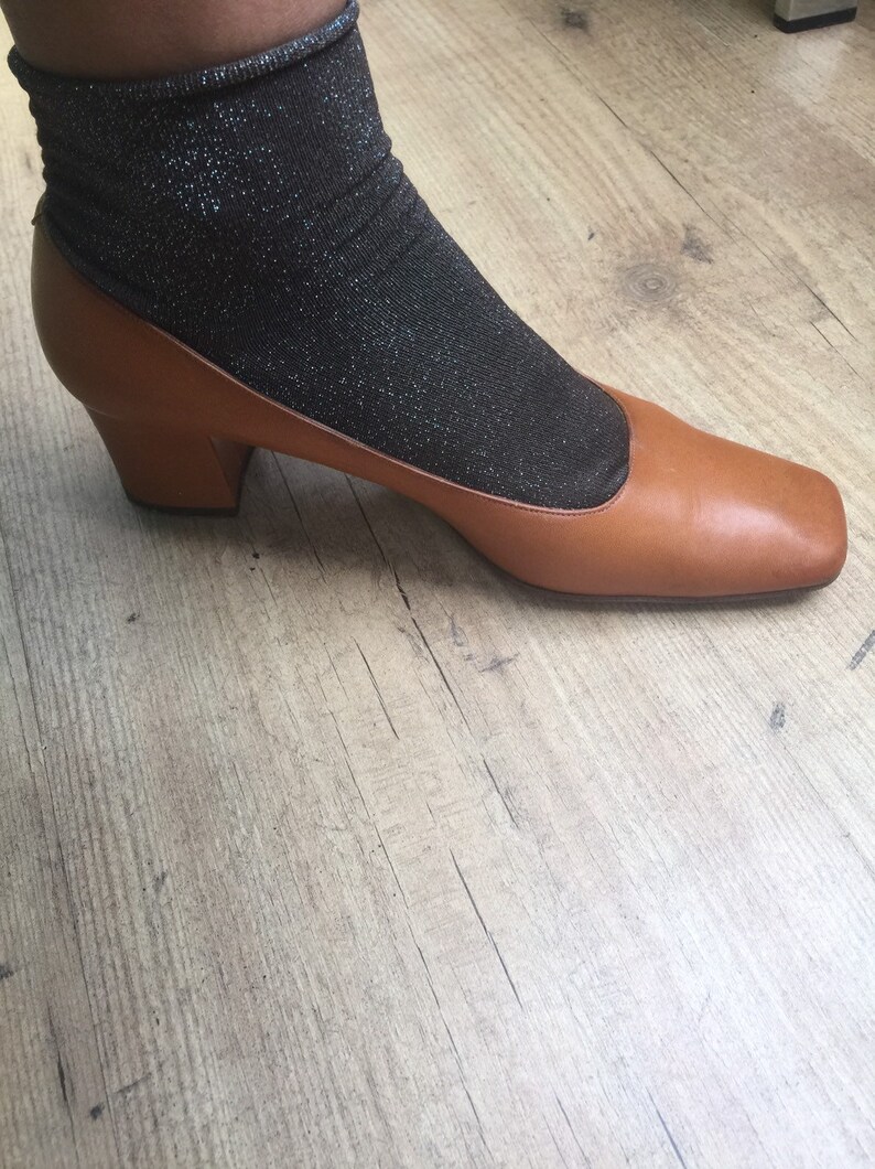 May include: A brown leather shoe with a square toe and a small heel. The shoe is paired with a black, sparkly sock. The shoe is on a light brown wooden floor.