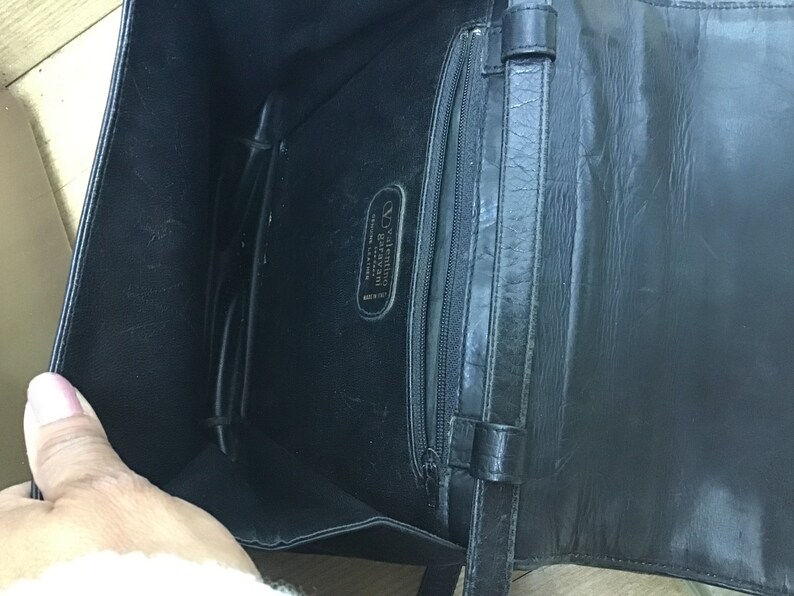 May include: Black leather handbag with an open interior, featuring a zippered pocket and a strap. The interior displays a label with the brand name "Valentino Garavani" and "Genuine Leather Made in Italy".
