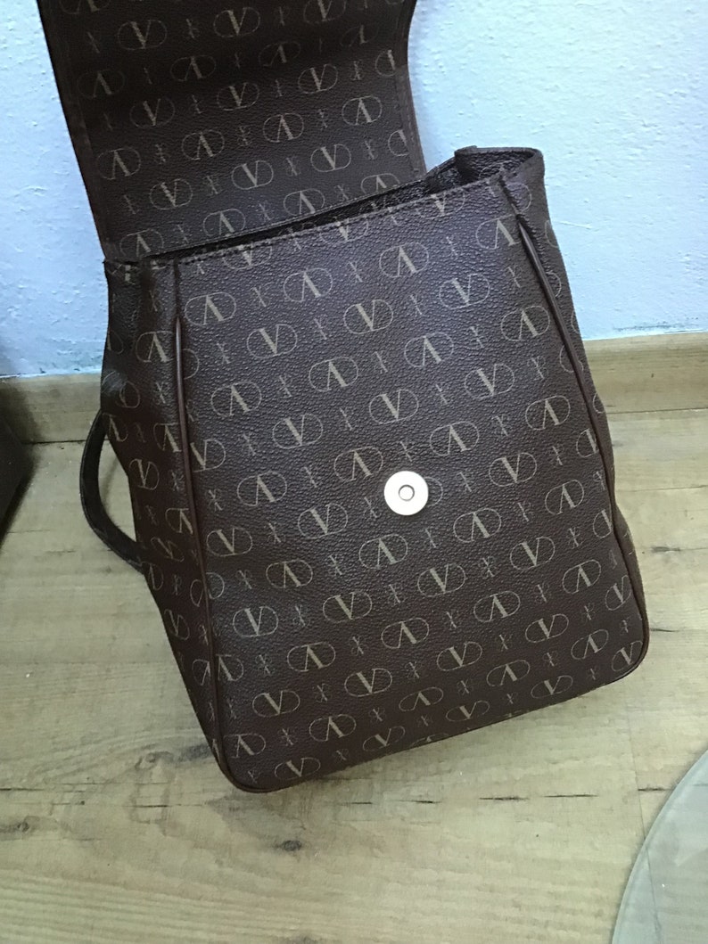 K&ouml;nnte beinhalten: Ein brauner Rucksack aus Kunstleder mit einem sich wiederholenden Muster des Buchstabens "V" in einer helleren braunen Farbe. Der Rucksack hat einen einzigen Druckknopfverschluss an der Vorderseite.