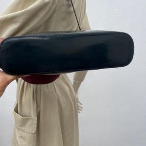 Puede incluir: Un bolso de hombro negro con un acabado liso y brillante y una base rectangular. El bolso cuenta con una sola correa fina unida a la parte superior, mostrando un dise&ntilde;o elegante con l&iacute;neas limpias y un color oscuro s&oacute;lido en todo el exterior.