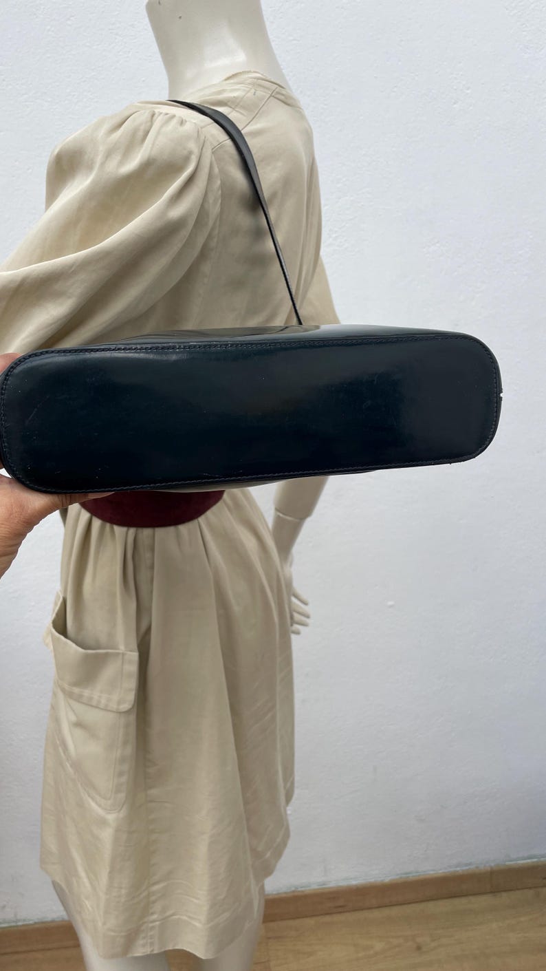Puede incluir: Un bolso de hombro negro con un acabado liso y brillante y una base rectangular. El bolso cuenta con una sola correa fina unida a la parte superior, mostrando un dise&ntilde;o elegante con l&iacute;neas limpias y un color oscuro s&oacute;lido en todo el exterior.