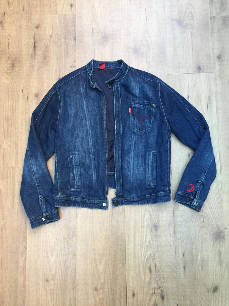 Peut inclure: Une veste en jean bleu avec une fermeture &eacute;clair et un logo Levi's rouge sur la poche. La veste a un col montant et des manches longues.