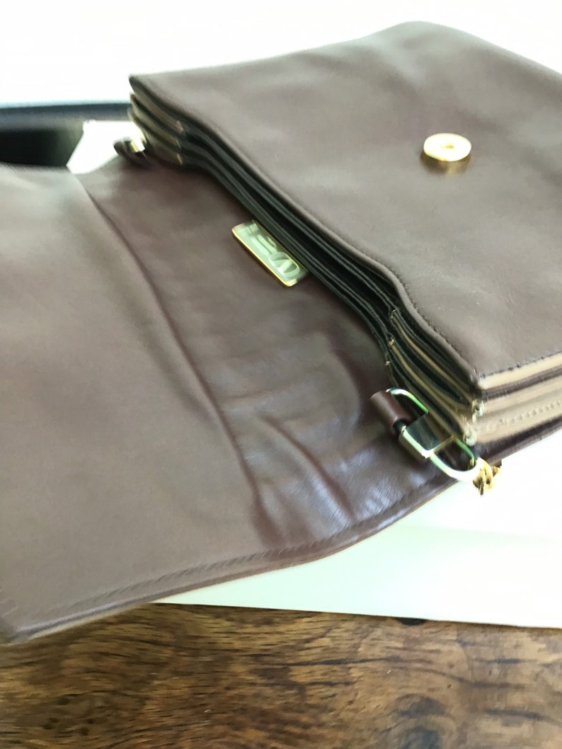 May include: A brown leather handbag with a gold-colored clasp and a gold-colored logo. The bag is open, revealing the interior and layered design. The bag has a magnetic closure.