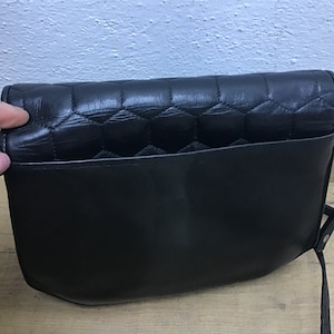 May include: Black leather shoulder bag with a quilted flap and a smooth body. The bag has a long strap and a pocket on the back. The bag is sitting on a wooden surface.