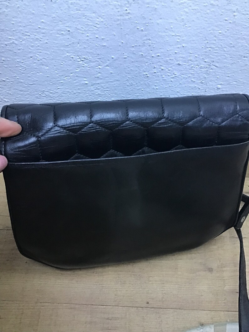 May include: Black leather shoulder bag with a quilted flap and a smooth body. The bag has a long strap and a pocket on the back. The bag is sitting on a wooden surface.