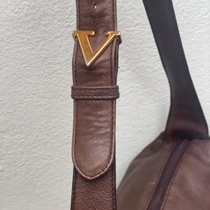 May include: Close-up of a brown leather shoulder bag with a gold-colored "V" buckle. The bag features a long strap and a zippered compartment, showcasing a classic design.