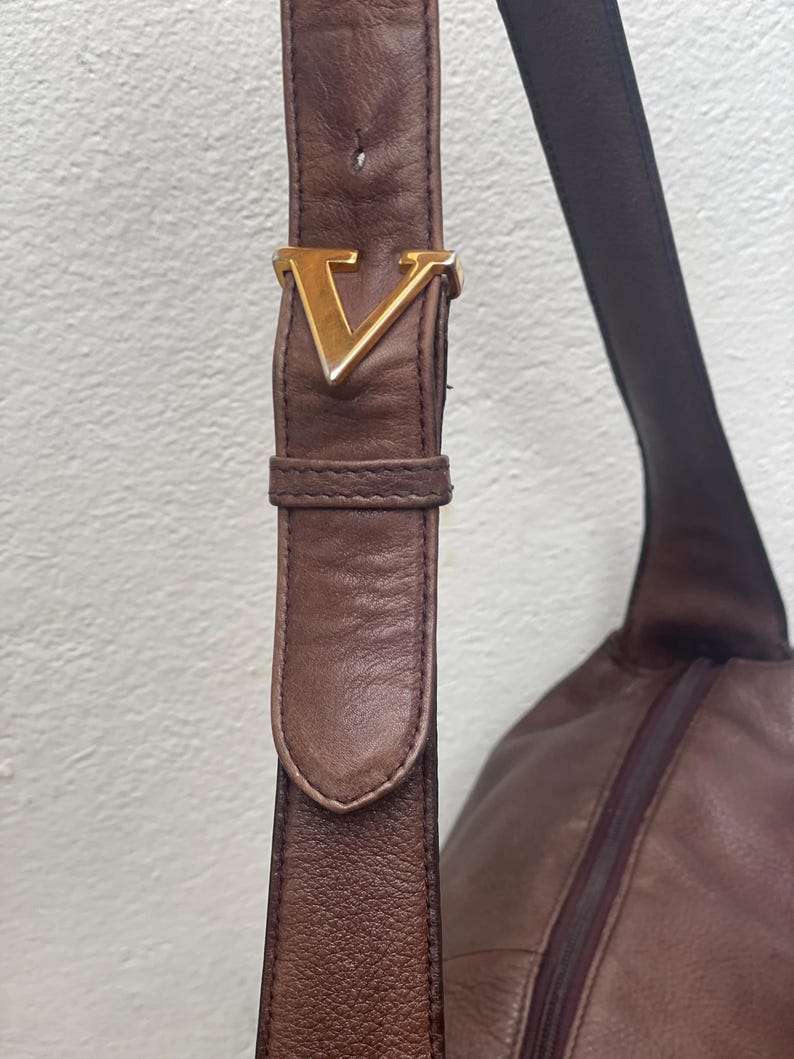 May include: Close-up of a brown leather shoulder bag with a gold-colored "V" buckle. The bag features a long strap and a zippered compartment, showcasing a classic design.