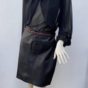 May include: A black outfit on a mannequin. The outfit includes a sheer black top, a black leather skirt, and a brown belt. The top has long sleeves and a high collar. The skirt is knee-length. The belt is made of woven leather.