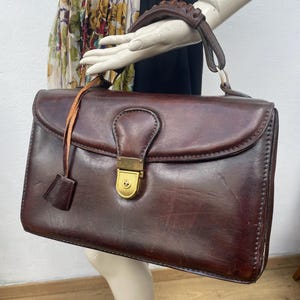 Op de afbeelding: Een vintage, rechthoekige bruinleren handtas met een klepsluiting en een goudkleurige sluiting. De tas heeft een handvat aan de bovenkant en een leren riem met een klein, decoratief leren stuk. De tas wordt op een mannequin getoond.