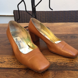 May include: A pair of brown leather pumps with square toes and block heels. The shoes have gold-colored insoles and the Valentino logo is visible inside. The shoes are displayed on a wooden surface.