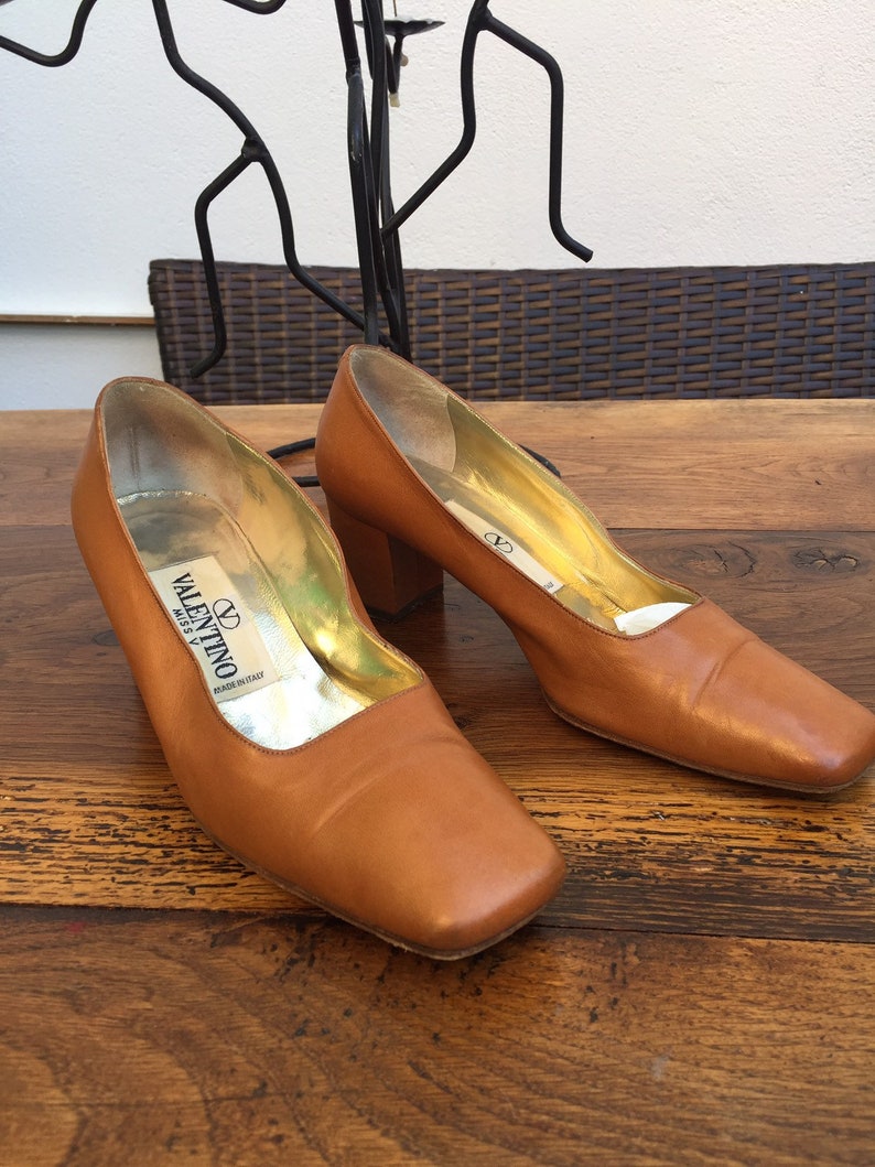 May include: A pair of brown leather pumps with square toes and block heels. The shoes have gold-colored insoles and the Valentino logo is visible inside. The shoes are displayed on a wooden surface.
