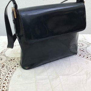 Puede incluir: Un bolso de hombro de charol negro con cierre de solapa rectangular, acabado brillante y liso, y herrajes de metal dorado que conectan la correa con el cuerpo principal del bolso, presentando una silueta estructurada.