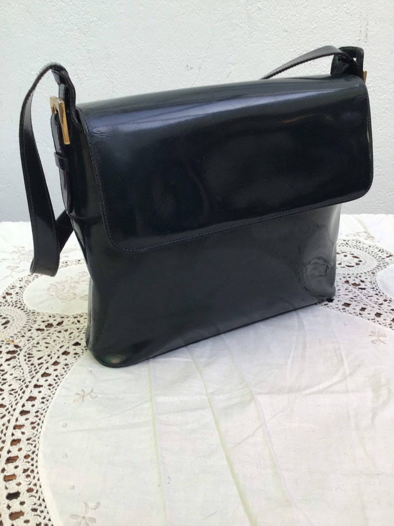 Puede incluir: Un bolso de hombro de charol negro con cierre de solapa rectangular, acabado brillante y liso, y herrajes de metal dorado que conectan la correa con el cuerpo principal del bolso, presentando una silueta estructurada.
