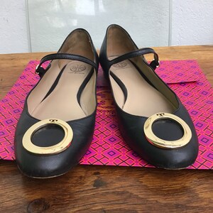 May include: A pair of black leather ballet flats with a gold-tone ring detail on the toe. The flats have a strap across the top of the foot and are displayed on a patterned surface.