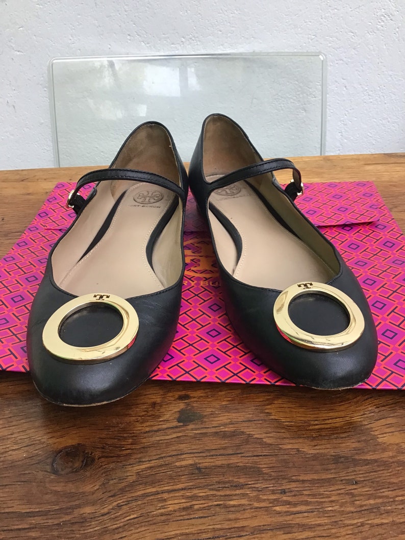 May include: A pair of black leather ballet flats with a gold-tone ring detail on the toe. The flats have a strap across the top of the foot and are displayed on a patterned surface.