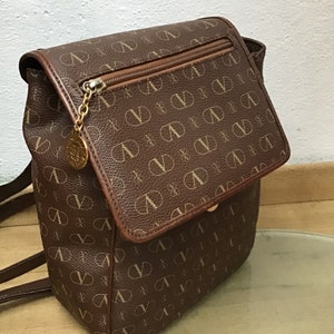 K&ouml;nnte beinhalten: Ein brauner Lederrucksack mit einem Monogrammbild. Der Rucksack hat einen Klappenverschluss mit einer Rei&szlig;verschlusstasche darunter. Die Tr&auml;ger sind verstellbar und der Rucksack hat einen goldfarbenen Rei&szlig;verschluss.