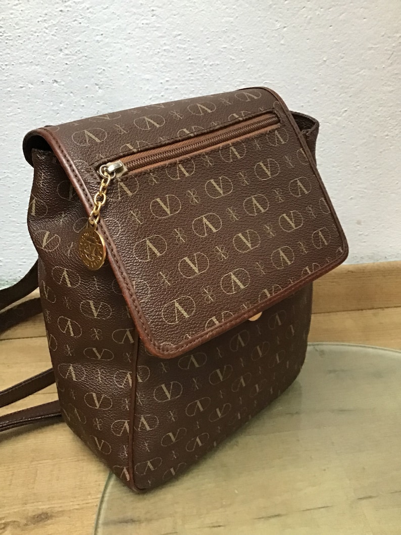 K&ouml;nnte beinhalten: Ein brauner Lederrucksack mit einem Monogrammbild. Der Rucksack hat einen Klappenverschluss mit einer Rei&szlig;verschlusstasche darunter. Die Tr&auml;ger sind verstellbar und der Rucksack hat einen goldfarbenen Rei&szlig;verschluss.