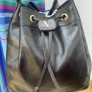 May include: Black leather bucket bag with a drawstring closure and gold-tone hardware. The bag features a textured finish and a small logo emblem on the front. The bag has a shoulder strap and a drawstring closure.