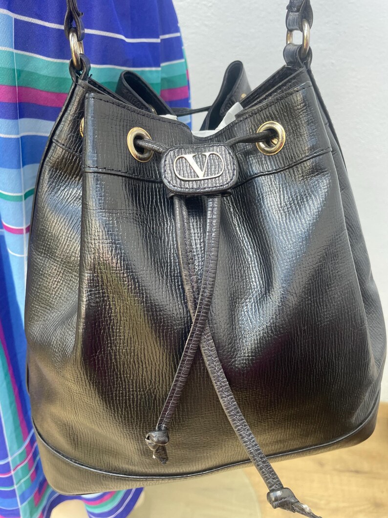 May include: Black leather bucket bag with a drawstring closure and gold-tone hardware. The bag features a textured finish and a small logo emblem on the front. The bag has a shoulder strap and a drawstring closure.