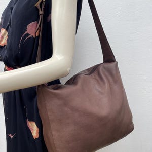 May include: A brown leather shoulder bag with a suede strap. The bag has a soft, slouchy design and a zipper closure. The bag is being held by a mannequin arm, and the background is a plain white wall.