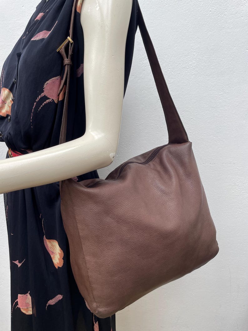May include: A brown leather shoulder bag with a suede strap. The bag has a soft, slouchy design and a zipper closure. The bag is being held by a mannequin arm, and the background is a plain white wall.