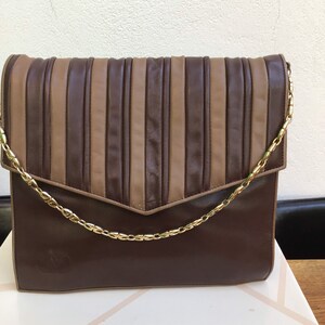 May include: A brown leather clutch with a gold chain strap. The flap features vertical pleated stripes in alternating shades of brown and tan. The bag has a rectangular shape and a subtle logo on the front.