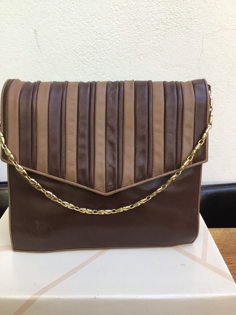 May include: A brown leather clutch with a gold chain strap. The flap features vertical pleated stripes in alternating shades of brown and tan. The bag has a rectangular shape and a subtle logo on the front.