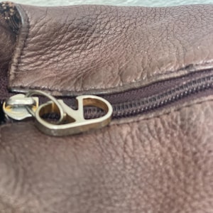 May include: Close-up of a brown leather item with a zipper. The zipper pull is silver-colored and features a stylized letter "V" and "D". The leather has a textured surface.