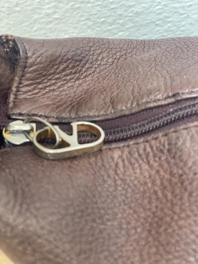 May include: Close-up of a brown leather item with a zipper. The zipper pull is silver-colored and features a stylized letter "V" and "D". The leather has a textured surface.