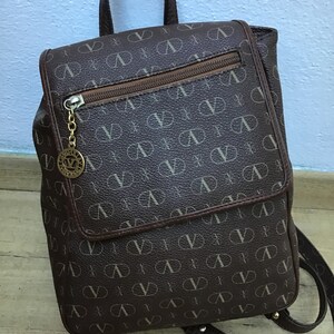 K&ouml;nnte beinhalten: Ein brauner Lederrucksack mit einem Monogramm-Muster des Buchstabens "V". Der Rucksack hat einen oberen Griff, verstellbare Tr&auml;ger und eine Rei&szlig;verschlusstasche auf der Vorderseite.