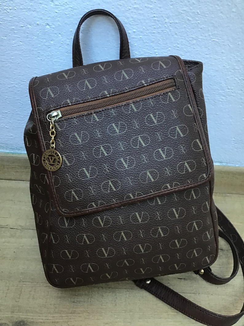 K&ouml;nnte beinhalten: Ein brauner Lederrucksack mit einem Monogramm-Muster des Buchstabens "V". Der Rucksack hat einen oberen Griff, verstellbare Tr&auml;ger und eine Rei&szlig;verschlusstasche auf der Vorderseite.