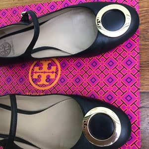 May include: Black leather ballet flats with a gold-tone circular buckle and ankle strap. The shoes are displayed on a pink and orange patterned box. The brand logo is visible on the buckle and insole.