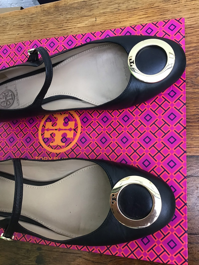 May include: Black leather ballet flats with a gold-tone circular buckle and ankle strap. The shoes are displayed on a pink and orange patterned box. The brand logo is visible on the buckle and insole.