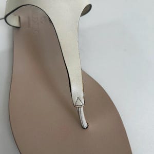 May include: Close-up of a white leather sandal with a thong strap design. The sandal features a light brown footbed and a metallic gold pyramid stud. The brand name "VALENTINO" is visible on the side.