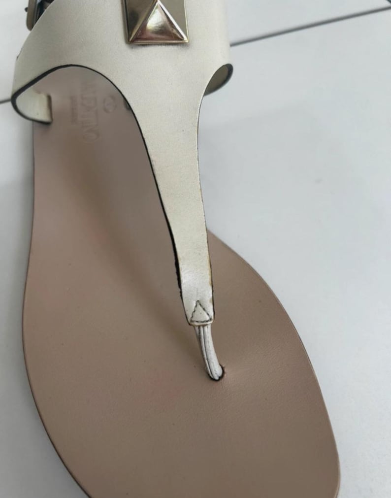 May include: Close-up of a white leather sandal with a thong strap design. The sandal features a light brown footbed and a metallic gold pyramid stud. The brand name "VALENTINO" is visible on the side.