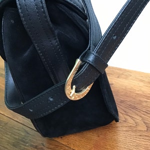 May include: Black handbag with a suede body and leather straps. The bag features a gold-colored buckle on the strap. The bag is set against a wooden surface.