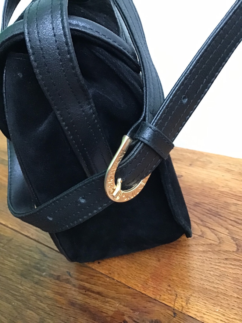 May include: Black handbag with a suede body and leather straps. The bag features a gold-colored buckle on the strap. The bag is set against a wooden surface.