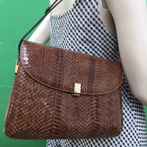 Può includere: Una borsa a tracolla in pelle di serpente marrone con motivo a scaglie testurizzato, patta ripiegabile e chiusura rettangolare in metallo dorato con inserto centrale bianco. La borsa presenta una sottile tracolla in pelle di serpente abbinata.