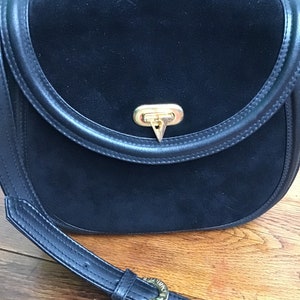May include: Black suede crossbody bag with a curved flap and a gold-tone clasp. The bag features a black leather strap with a gold buckle. The bag's design is classic and elegant.