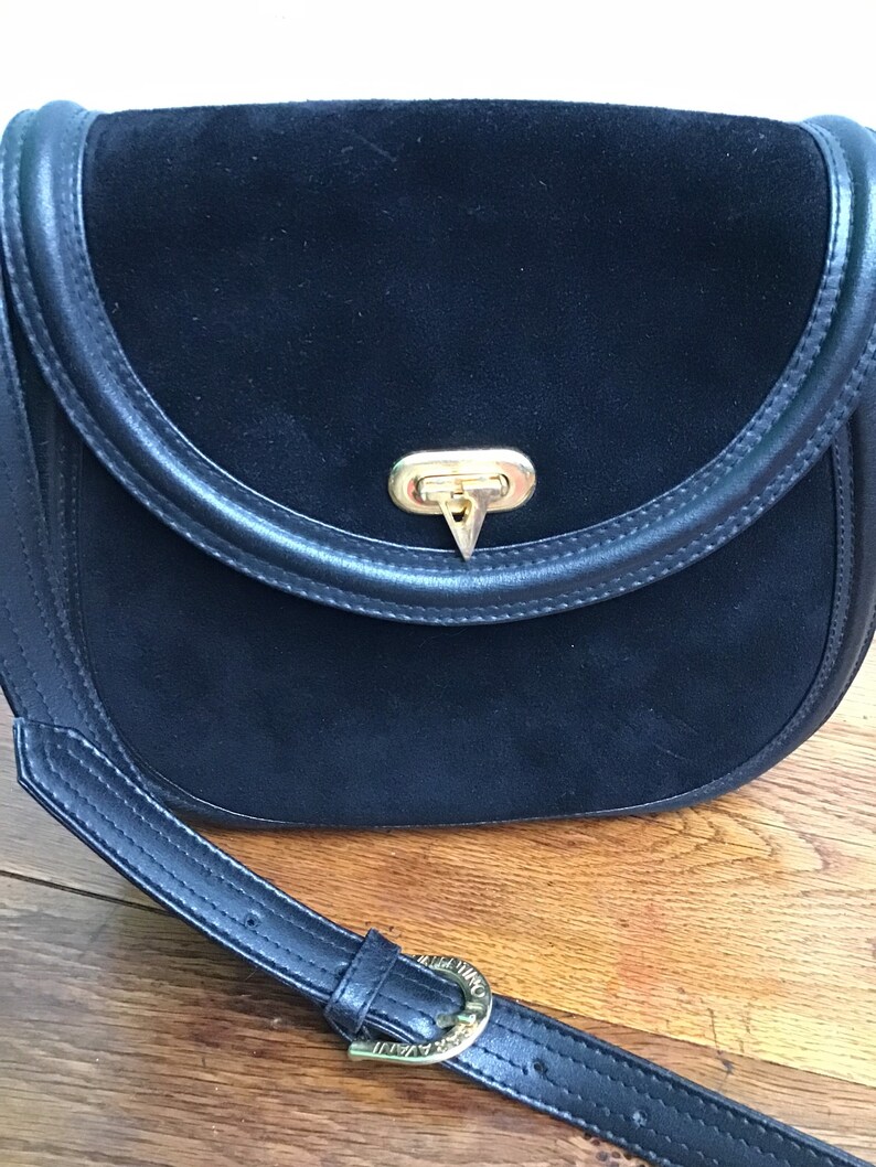 May include: Black suede crossbody bag with a curved flap and a gold-tone clasp. The bag features a black leather strap with a gold buckle. The bag's design is classic and elegant.
