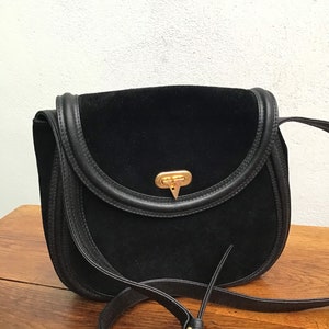 May include: Black suede crossbody bag with a rounded flap and leather trim. The bag features a gold-tone clasp and a long, adjustable strap. The bag is sitting on a wooden surface.