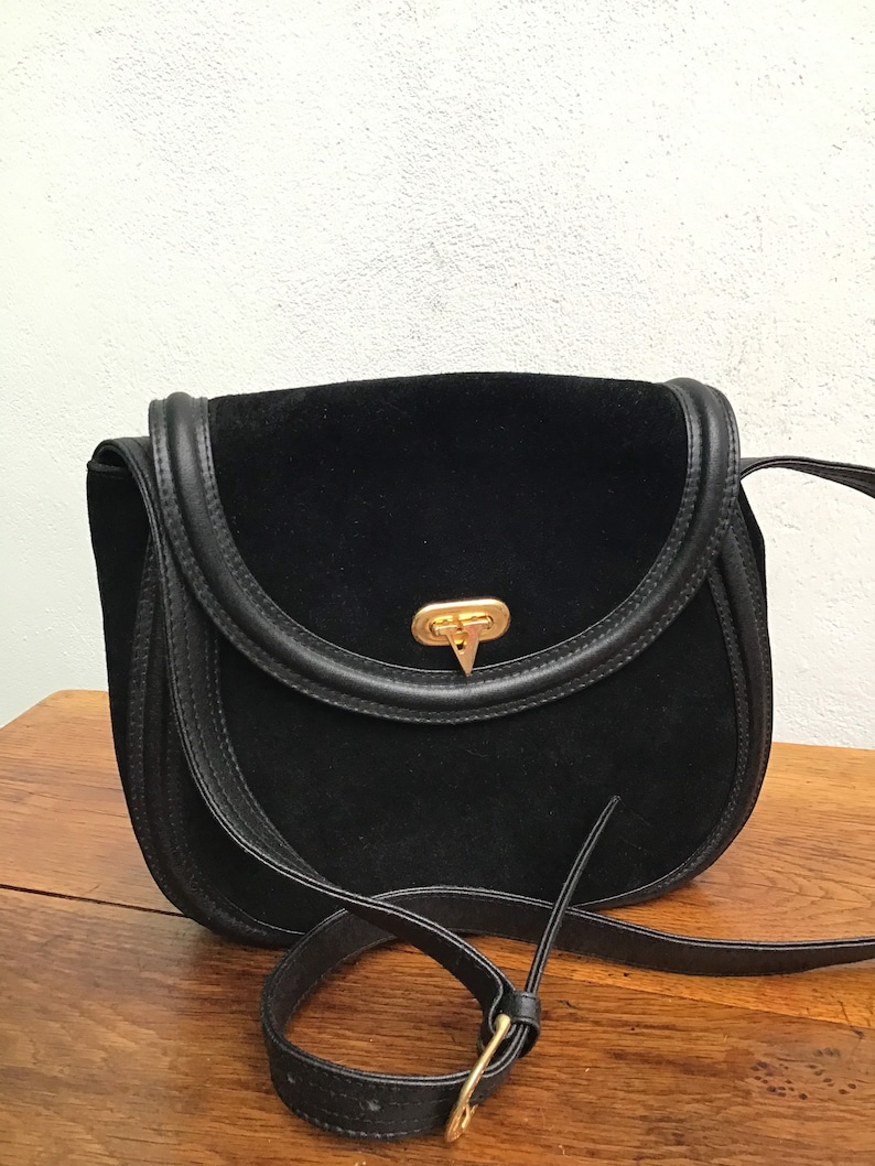 May include: Black suede crossbody bag with a rounded flap and leather trim. The bag features a gold-tone clasp and a long, adjustable strap. The bag is sitting on a wooden surface.