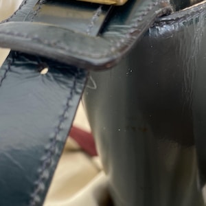 Puede incluir: Un bolso de mano de charol negro con acabado brillante y una correa estructurada. La correa est&aacute; asegurada con una hebilla rectangular de metal dorado que muestra un peque&ntilde;o logotipo V grabado en la parte frontal, con costuras visibles.