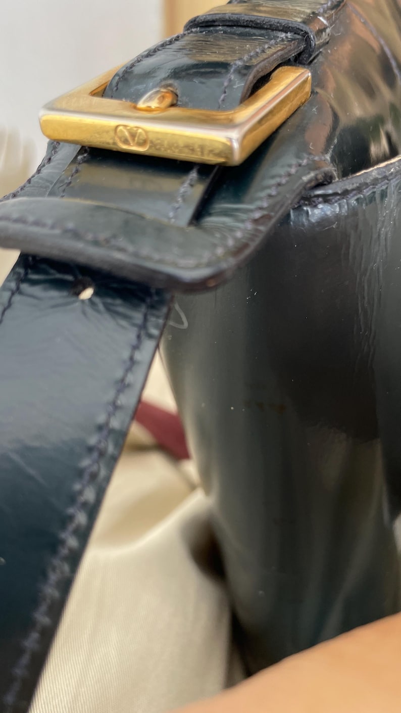 Puede incluir: Un bolso de mano de charol negro con acabado brillante y una correa estructurada. La correa est&aacute; asegurada con una hebilla rectangular de metal dorado que muestra un peque&ntilde;o logotipo V grabado en la parte frontal, con costuras visibles.