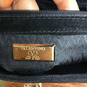 May include: Close-up of a black handbag interior with a gold-colored metal plaque. The plaque reads "VALENTINO LES SACS MADE IN ITALY". The bag has a zipper and a gold-colored clasp.